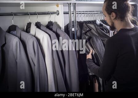 Portrait d'un jeune gars barbu millénaires. Dans un magasin de vêtements pour hommes, un gars au milieu d'un comptoir avec des vestes d'affaires choisit un cuir brutal Banque D'Images