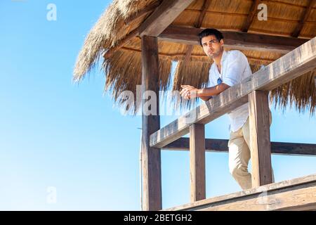 L'homme se trouve sur un bungalow en bois à la plage près de la mer. Élégant et beau jeune homme avec une chemise blanche dans une cabane en bois. Vacances d'été par la mer. Banque D'Images