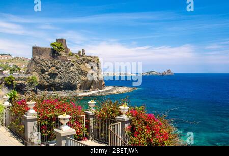 Ancien château médiéval de Norman ACI Castello avec murs en pierre sur la côte rocheuse près de l'eau de la mer méditerranée et clôture avec fleurs rouges en premier plan, Catane, Banque D'Images