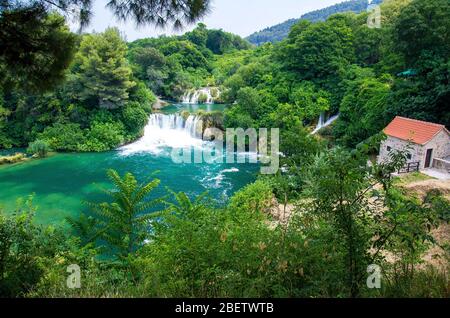 Belles cascades fluviales parmi les plantes vertes, les arbres, les forêts et le bâtiment de moulin à pierre avec toit en tuiles orange dans le parc national de Krka, Dalmatie, Croatie, Banque D'Images