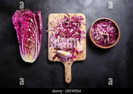 Chou rouge chinois haché (wombok violet) sur planche à découper en bois et bol sur table en pierre. Moitié de chou rouge Napa sur fond noir, vue de dessus. Cuire Banque D'Images