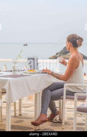Une femme buvant un café avec son ordinateur portable assis dans un restaurant près de la plage tôt le matin. Banque D'Images