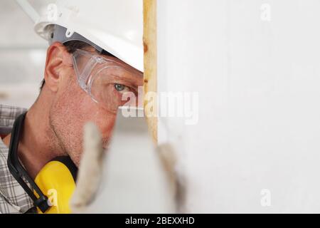 homme travailleur mesure l'angle du mur avec un casque carré, des lunettes et des écouteurs de protection auditive, sur le chantier de construction intérieur Banque D'Images