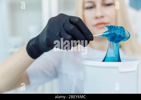 Maître de la cire. Préparation de la procédure d'épilation. Une fille blonde dans une robe médicale prend une cire bleue. Banque D'Images