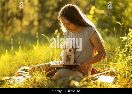 Une fille assise près d'un arbre et lisant un livre, tenant un chiot du labrador. Banque D'Images