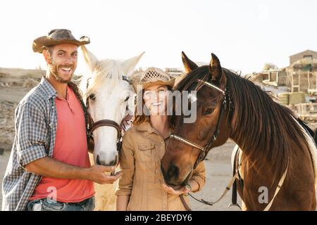 Heureux couple ayant plaisir avec les chevaux à l'intérieur stable - jeunes agriculteurs partageant le temps avec les animaux dans le ranch corral Banque D'Images