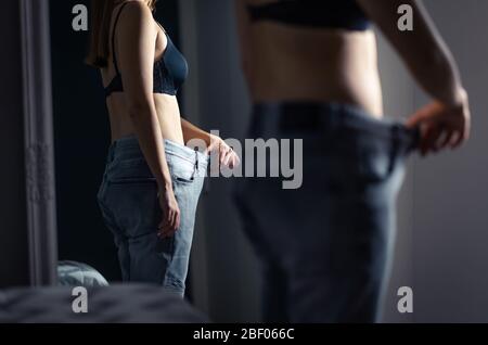 Petite femme mince portant un grand pantalon devant un miroir après perte de poids et régime. Fille sous-poids avec stress malsain d'être graisse. Banque D'Images