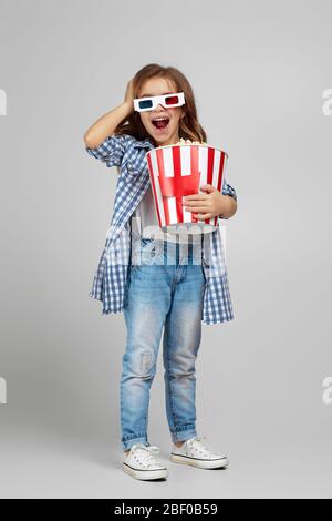 vue sur toute la longueur de la jolie fille caucasienne, agréablement surprise, portant des lunettes tridimensionnelles rouges et mangeant du pop-corn à partir d'un seau isolé sur fond gris. Banque D'Images