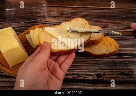 Quelqu'un répand le beurre sur des toasts de blé frais avec un couteau et un plat de beurre en bois avec un morceau de beurre et des tranches de fromage sur un fond en bois. Banque D'Images