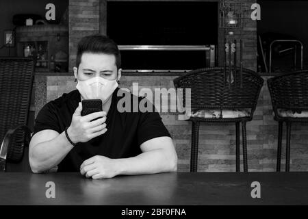 Homme portant un masque blanc pendant la quarantaine à la maison, en vérifiant les nouvelles sur le téléphone. Banque D'Images