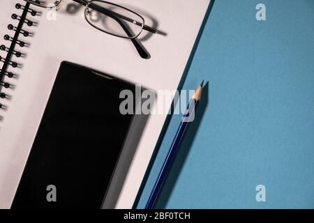 vue de dessus du téléphone portable noir, des crayons et du bloc-notes scolaire, des lunettes et du bloc-notes en spirale sur une table. Vue en grand angle du planificateur vide. Banque D'Images
