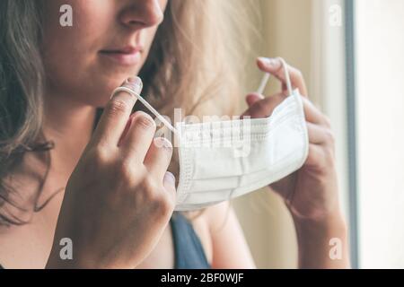 Photo détaillée d'une femme blanche non reconnaissable mettant sur un masque médical blanc pour prévenir l'infection. Fond flou, foyer sur le masque. Coronavirus, COVID-19 quarantaine. Concept de verrouillage. Banque D'Images