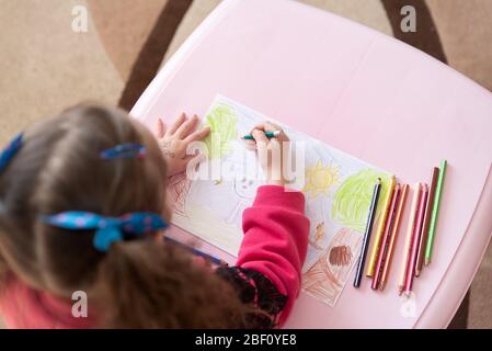 Drohobych, Ukraine - 16 avril 2020: La petite fille dessine une photo couleur sur un livre blanc, vue arrière d'en haut. Des crayons de couleur se trouvent sur la table. Thèmes Banque D'Images