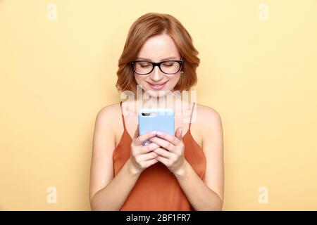 Studio portrait de jeune femme souriante portant des lunettes, regarde et écrit un message sur smartphone à son petit ami ou de vérifier les médias sociaux, isolat Banque D'Images