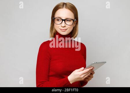 Portrait d'affaires d'une femme élégante et confiante en col de cygne rouge, portant des lunettes optiques, tenant une tablette informatique, en restant isolée sur le gris b. Banque D'Images
