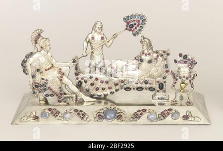 Groupe sculptural. Argent moulé, un tableau incrusté de bijoux montrant un soldat romain en pleine régalia assis face à la femme égyptienne se reposant sur le canapé, et bronzé par un serviteur debout; petite table de trépied à droite du canapé, tenant un bol de fruits ou de fleurs et urne sur le plateau en dessous. Banque D'Images