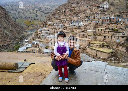 Palangan, Kurdistan iranien - 15 novembre 2013: Portrait de la petite fille kurde et mignonne huged par son père tout en posant avec le village de pierre de Palangan en arrière-plan Banque D'Images