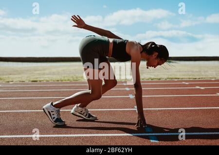 Vue latérale du coureur féminin se préparer à la ligne de départ sur la piste de course au stade. Femme athlète prenant position sur ses marques pour commencer le ru Banque D'Images