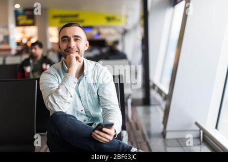 Il envoie un SMS très bien. Portrait de beau homme dans un port décontracté tenant ses bagages et la messagerie par son téléphone mobile tout en étant assis dans le hall du TH Banque D'Images