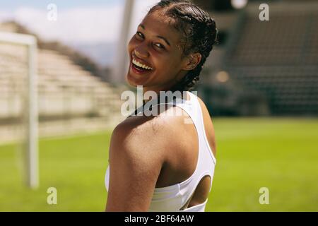 Gros plan d'une athlète féminine qui regarde son épaule et qui souriait. Femme dans des vêtements de sport regardant l'appareil photo et souriant sur le terrain de sport. Banque D'Images