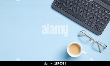 Table de bureau avec clavier d'ordinateur portable, lunettes, tasse à café sur fond bleu. Vue sur le dessus bureau moderne, espace de travail féminin avec espace de copie. Banque D'Images