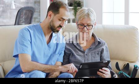 Assistant masculin enseignant aux aînés retraités âgés comment utiliser son ordinateur tablette dans la maison de soins pour sa thérapie Banque D'Images
