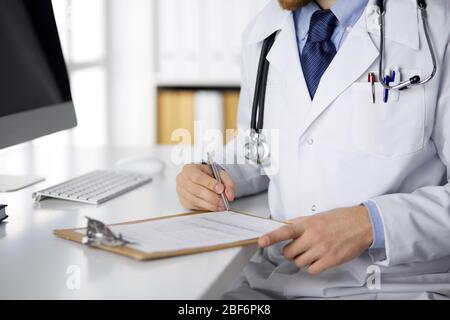 Sympathique médecin barbu rouge assis et écrire avec presse-papiers dans la clinique, concept de médecine rapprochée Banque D'Images