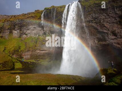 Un arc-en-ciel provoqué par le jet de la cascade Seljalandsfoss dans le sud de l'Islande, avec un touriste qui la répand au premier plan Banque D'Images