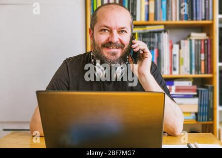 Concept de travail à domicile. Homme barbu adulte concentré, travaillant en ligne de la maison sur ordinateur portable, réserver des étagères derrière lui Banque D'Images