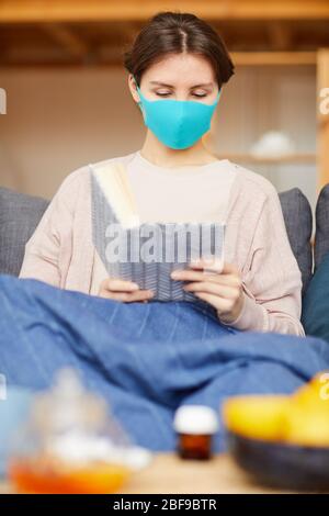 Jeune femme malade portant un masque de protection assis sur un canapé et lisant un livre à la maison Banque D'Images
