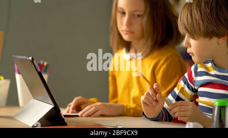 Étudiants à distance à la maison. La sœur plus âgée contrôle les devoirs du jeune frère. Au premier plan, un garçon tient un crayon dans sa main. Banque D'Images