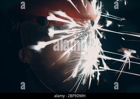 Homme barbu adulte dans un masque de protection transparent avec des particules métalliques volantes étincelles dans l'obscurité Banque D'Images