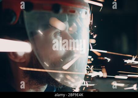 Homme barbu adulte dans un masque de protection transparent avec des particules métalliques volantes étincelles dans l'obscurité Banque D'Images