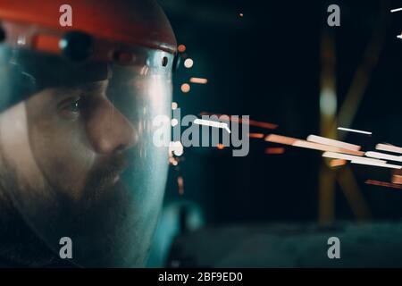 Homme barbu adulte dans un masque de protection transparent avec des particules métalliques volantes étincelles dans l'obscurité Banque D'Images