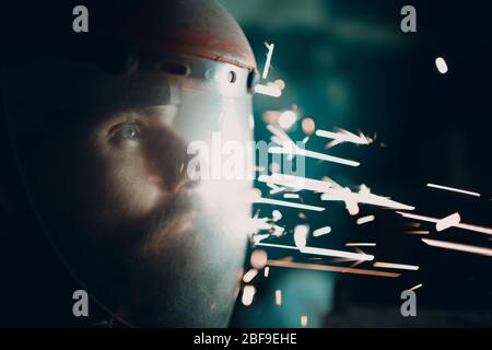 Homme barbu adulte dans un masque de protection transparent et une scie à moulin avec des particules de métal volants étincelles dans l'obscurité Banque D'Images