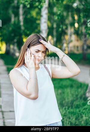 Jeune femme avec de longs cheveux dans un t-shirt blanc vêtu de parler sur le téléphone mobile et a mal de tête, les mains sont sur sa tête Banque D'Images