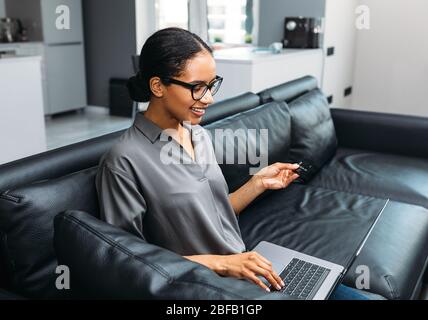 Jeune femme gaie utilisant une carte de crédit et un ordinateur portable pour faire des achats en ligne à partir de la maison Banque D'Images