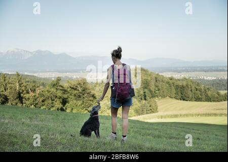 Vue de derrière une jeune femme randonneur, debout dans un pré vert avec son chien noir à côté d'elle, en profitant de la vue. Banque D'Images