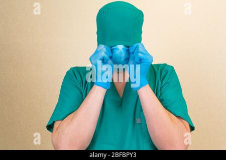 Médecin en costume vert et gants bleus se cachant le visage sous masque un chapeau isolé Banque D'Images