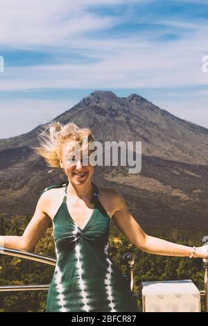 Jeune blonde femme avec robe verte debout devant le volcan du Mont Batur sur l'île de Bali pendant les jours nuageux et venteux Banque D'Images