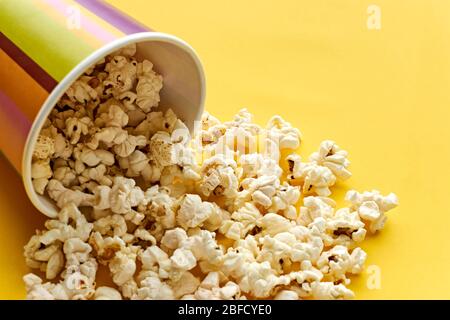 Popcorn dans un gobelet à papier. Seau à gobelets en papier rayé multicolore avec pop-corn renversé sur fond jaune avec espace de copie. Concept cinéma ou cinéma. Banque D'Images