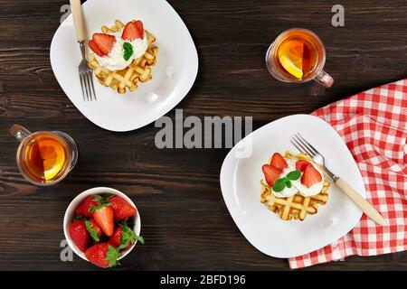Le petit déjeuner est magnifique. Gaufres belges viennois décorées de crème et de fraises, thé et serviette à carreaux sur un fond rustique en bois sombre. Banque D'Images
