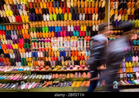 Les piétons se promènent devant un stand de rue vendant des chaussons en cuir traditionnels colorés dans la médina (vieux quartier) de Fez, au Maroc. Banque D'Images