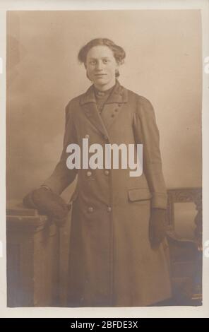 Carte postale de studio photographique vintage montrant une dame portant une couche de recouvrement, des gants et tenant un chapeau de laine. Banque D'Images