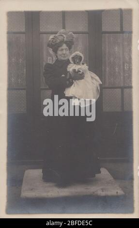 Vintage Edwardian 1910 carte postale photographique d'une dame habilement vêtue tenant un petit chien dans ses bras. Le chien porte un modèle et un capot Banque D'Images