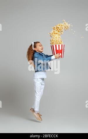 vue sur toute la longueur d'une fille caucasienne souriante avec des lunettes tridimensionnelles rouges, qui tient un seau avec du pop-corn volant et qui sautait sur fond gris. Banque D'Images