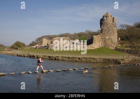 Château d'Ogmore avec une jeune femme traversant la rivière Ewenny sur les pierres de pas avec son chien Banque D'Images