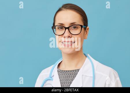 Portrait de jeune femme médecin dans des lunettes et dans un manteau blanc souriant à l'appareil photo sur fond bleu Banque D'Images