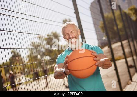 Jouer au basket-ball. Homme d'âge moyen et gai dans des vêtements de sport tenant le ballon de basket-ball et souriant à l'appareil photo tout en se tenant sur un terrain de basket-ball extérieur Banque D'Images