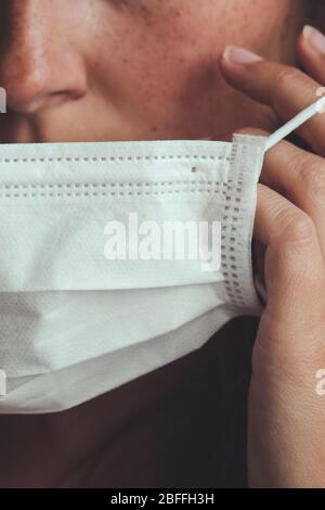 Photo détaillée d'une femme blanche non reconnaissable mettant sur un masque médical blanc pour empêcher la grippe. Fond flou, foyer sur le masque. Coronavirus, COVID-19 quarantaine. Concept de verrouillage. Banque D'Images